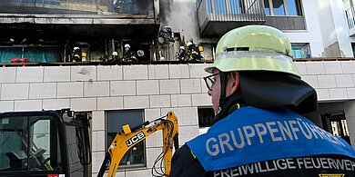 Feuerwehrmann mit Helm und 'Gruppenführer Freiwillige Feuerwehr München' auf der Jacke vor einem ausgebrannten Wohngebäude mit Feuerwehrleuten auf dem Balkon