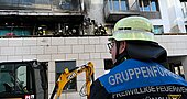 Feuerwehrmann mit Helm und 'Gruppenführer Freiwillige Feuerwehr München' auf der Jacke vor einem ausgebrannten Wohngebäude mit Feuerwehrleuten auf dem Balkon