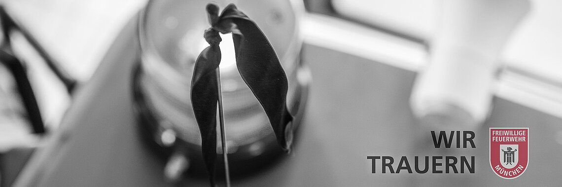 Trauerflor an einem Feuerwehrfahrzeug vor unscharfem Hintergrund mit Text 'WIR TRAUERN' und Logo der Freiwilligen Feuerwehr München