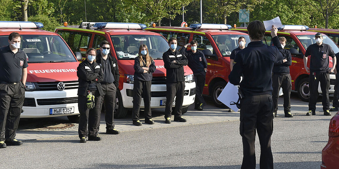 Feuerwehrleute in schwarzer Uniform und Mundschutz stehen vor Feuerwehrfahrzeugen bei einer Masken-Verteilaktion im Freien
