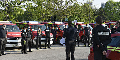 Feuerwehrleute in schwarzer Uniform und Mundschutz stehen vor Feuerwehrfahrzeugen bei einer Masken-Verteilaktion im Freien