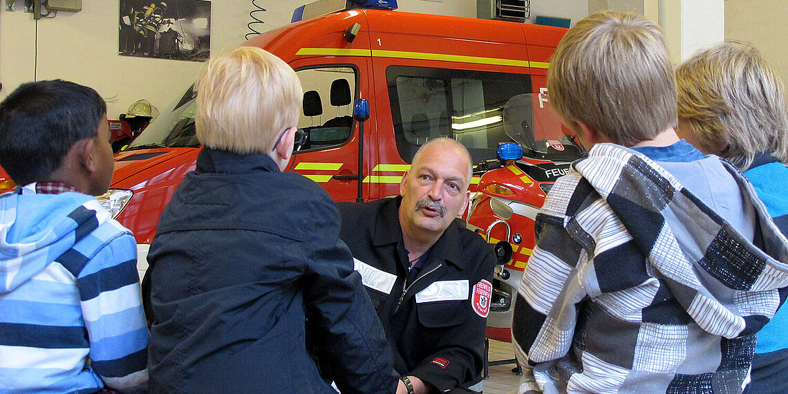 Feuerwehrmann erklärt Kindern vor einem Feuerwehrauto Brandschutzthemen