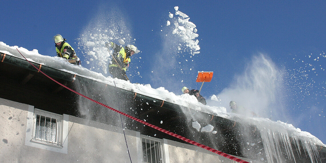 Mehrere Feuerwehrleute räumen Schnee mit Schaufeln von einem Dach, Schnee fliegt in die Luft, Szene bei klarem Himmel Mehrere Feuerwehrleute räumen Schnee mit Schaufeln von einem Dach, Schnee fliegt in die Luft, Szene bei klarem Himmel
