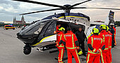 Polizeihubschrauber Airbus H145 auf der Theresienwiese, umgeben von Flughelfern in leuchtend orangen Schutzanzügen und Helmen, Einsatzfahrzeug im Hintergrund