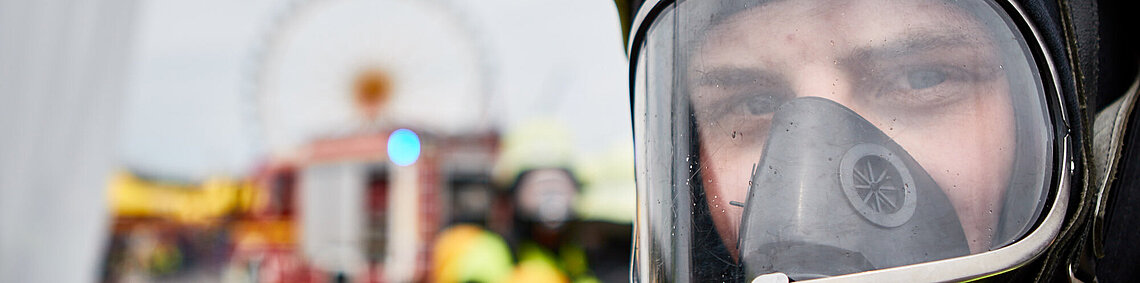 Nahaufnahme eines Feuerwehrmanns mit Atemschutzmaske und Helm, unscharfer Einsatzhintergrund mit Feuerwehrfahrzeug