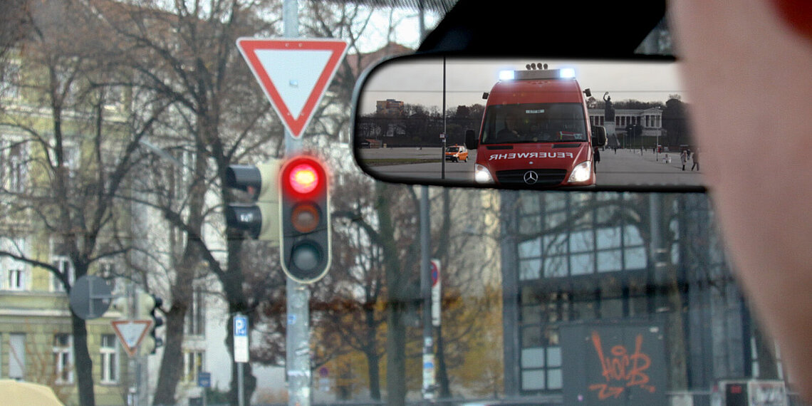 Im Rückspiegel zu sehen ist ein Einsatzfahrzeug der Feuerwehr mit Blaulicht, während die Ampel vor dem Fahrer rot zeigt Im Rückspiegel zu sehen ist ein Einsatzfahrzeug der Feuerwehr mit Blaulicht, während die Ampel vor dem Fahrer rot zeigt