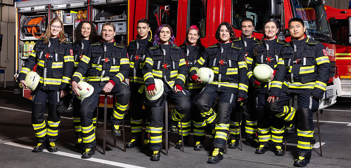 Neun Feuerwehrleute verschiedener Geschlechter und Herkunft in Schutzkleidung stehen vor einem Feuerwehrfahrzeug in der Fahrzeughalle, Teil der Kampagne DIVERSITÄT + INKLUSION