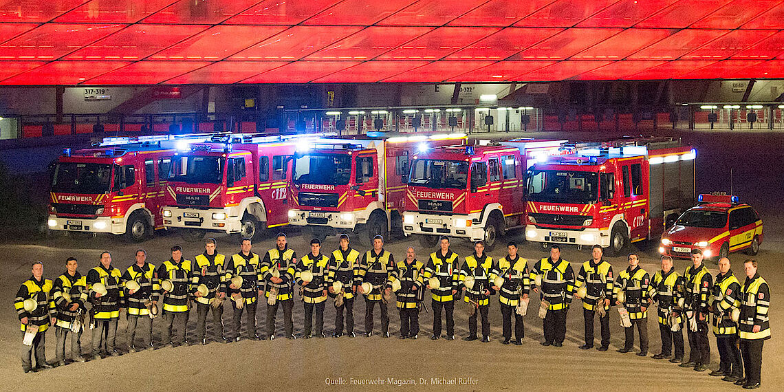 Mannschaft der Freiwilligen Feuerwehr München steht bei Dunkelheit in Schutzkleidung vor Einsatzfahrzeugen mit Blaulicht vor der Allianz Arena